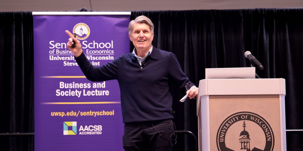 A speaker on stage near a podium and banner for the Sentry School of Business and Economics.