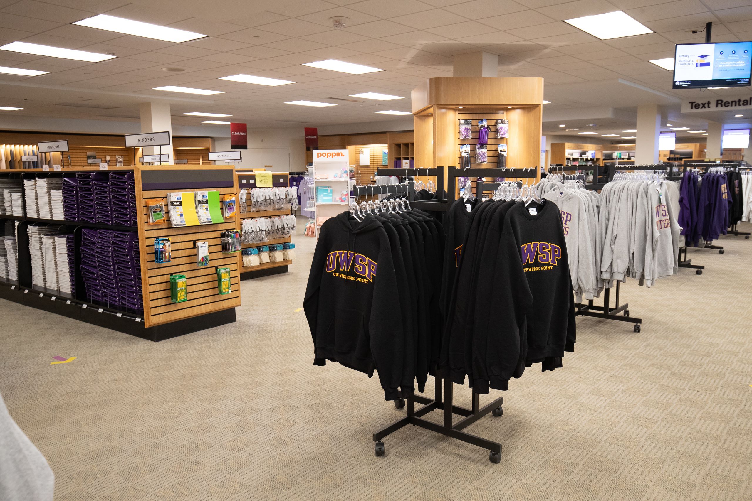 A spacious bookstore displays university merchandise, including UWSP hoodies, alongside shelves of stationery and school supplies.
