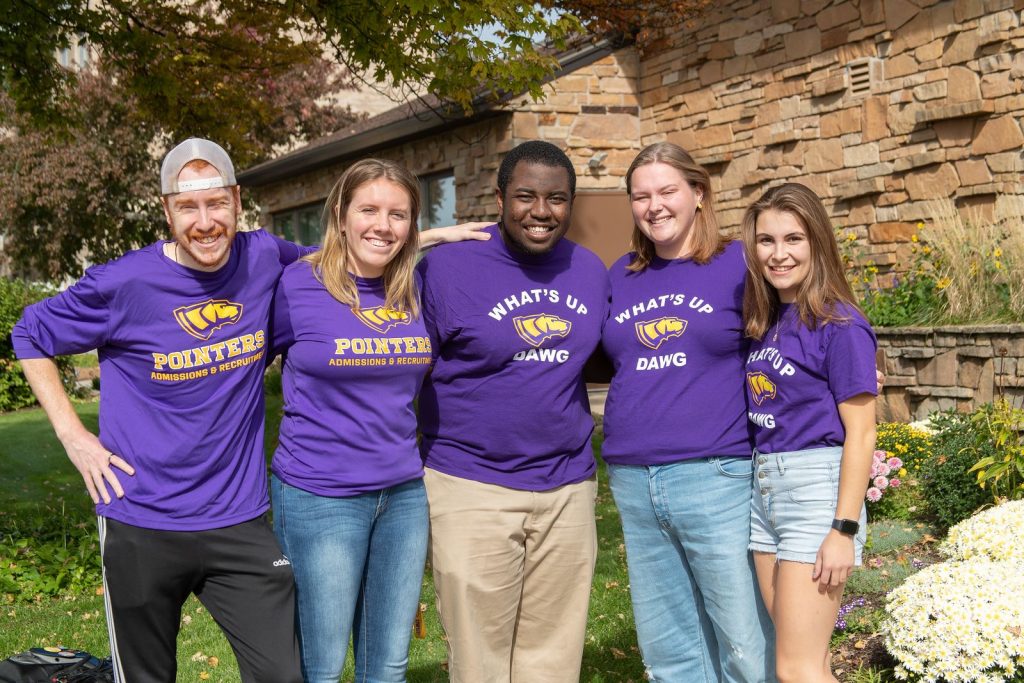 Five people are smiling and wearing matching purple shirts outdoors.