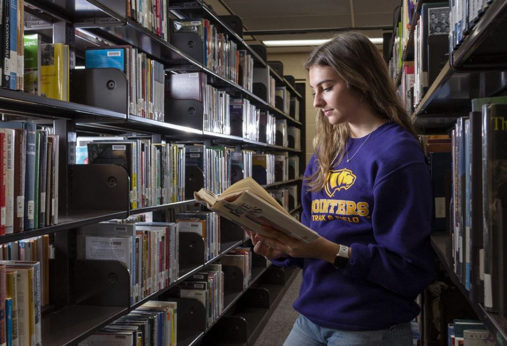 A student in a purple sweatshirt reads a book while standing between library bookshelves.